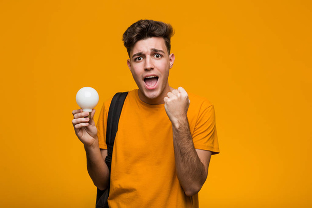 Young student man holding a light bulb celebrating a victory or success - Photo, Image
