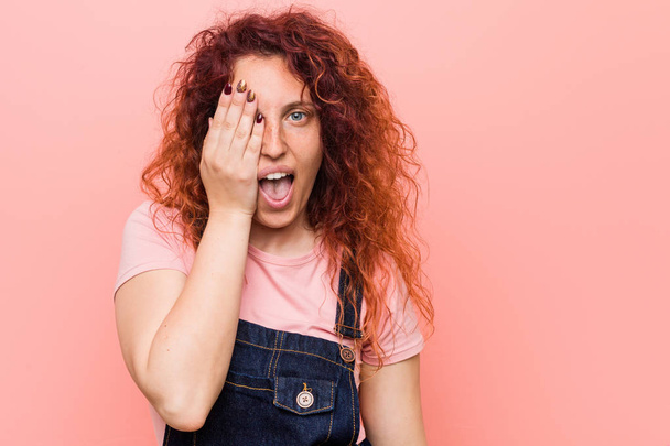 Young pretty ginger redhead woman wearing a jeans dungaree having fun covering half of face with palm. - Photo, Image