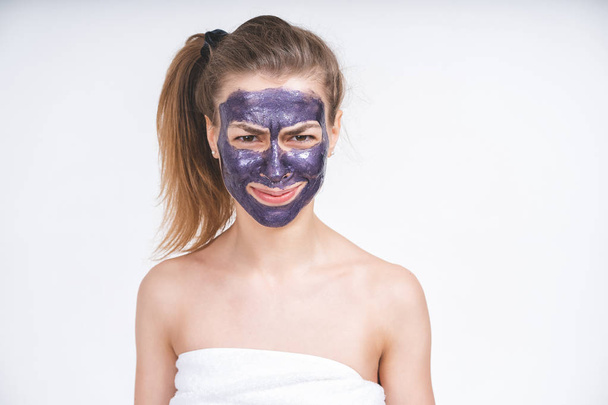 emotional portrait of a young beautiful european girl in violet cosmetic mask on her face. Wrapped in a towel. European, 22 years old, white, tail on the head, blank background, looking straight - Zdjęcie, obraz