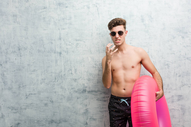 Young man holding a pink inflatable donut wearing a swimsuit upset screaming with tense hands. - 写真・画像