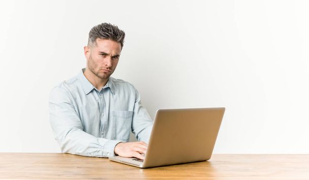 Young handsome man working with his laptop frowning face in displeasure, keeps arms folded. - 写真・画像