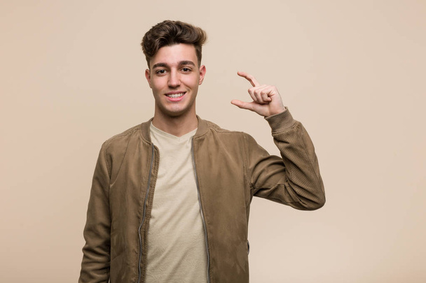 Jovem caucasiano vestindo uma jaqueta marrom segurando algo pouco com os dedos indicador, sorrindo e confiante
. - Foto, Imagem