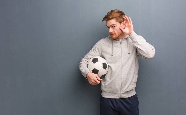 Молодой рыжий спортсмен пытается слушать сплетни. Он держит футбольный мяч.
. - Фото, изображение
