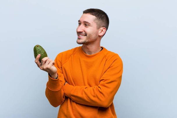 Jovem caucasiano segurando um abacate sorrindo confiante com braços cruzados
. - Foto, Imagem