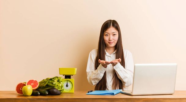 Jovem nutricionista mulher chinesa trabalhando com seu laptop segurando algo com palmas das mãos, oferecendo à câmera
. - Foto, Imagem