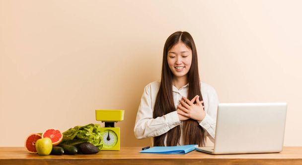 Jovem nutricionista mulher chinesa trabalhando com seu laptop rindo mantendo as mãos no coração, conceito de felicidade
. - Foto, Imagem