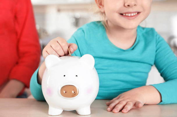 Cute girl putting coin into piggy bank at table, closeup. Saving money - Photo, Image