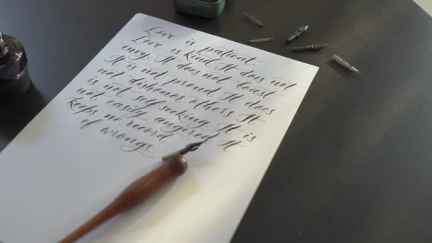 Close-up shot van een kalligrafiegereedschap en een fles inkt leggen rond een blad van een Witboek met verschillende regels uit een Bijbel over liefde is geschreven op het - Video