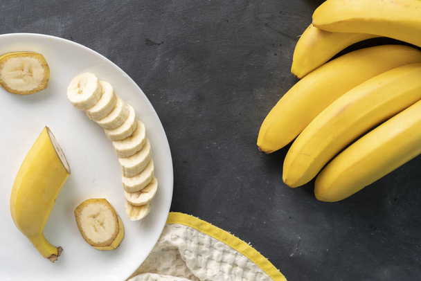 黒いテーブルの上の白いセラミックプレートに新鮮なスライスバナナ, 健康的な朝食のための食材を準備 - 写真・画像