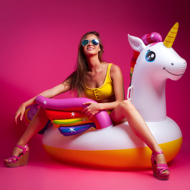 Konzept Sommerstimmung, Entspannung und Schönheit. Modernes Porträt einer jungen Frau in Shorts, Oberteil und Sandalen, die auf einer aufblasbaren Einhornmatratze auf isoliertem rosa Hintergrund ruht. - Foto, Bild