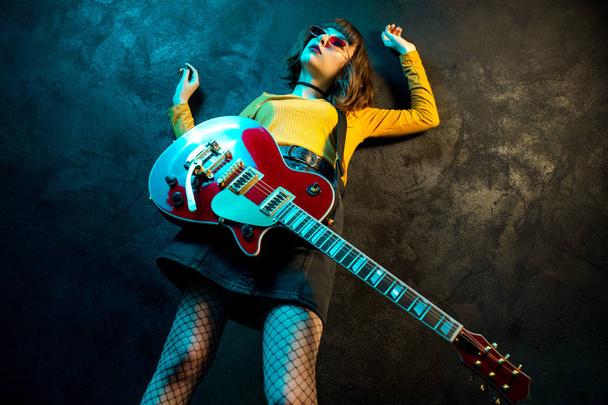 Neon ışıklar kırmızı gitar ile kıvırcık saçlı Moda genç hipster kadın. Rock müzisyeni elektrik gitar çalıyor. 90'ların stil konsepti. - Fotoğraf, Görsel