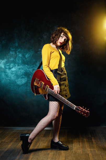 ネオンライトで赤いギターを持つ巻き毛を持つかなり若いヒップスターの女性。ロックミュージシャンがエレキギターを弾いている。90年代スタイルのコンセプト. - 写真・画像