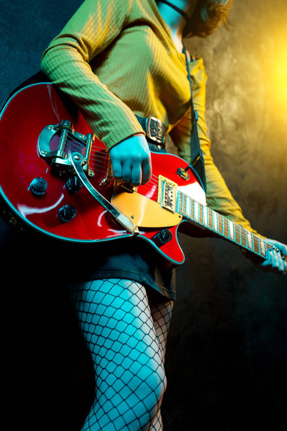 Close-up photo of young hipster woman legs and red guitar in neon lights. Rock musician is playing electrical guitar. 90s style concept. - Zdjęcie, obraz