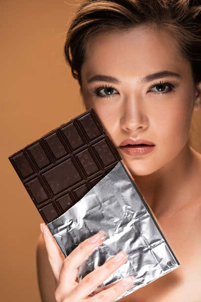 νεαρή γυμνή γυναίκα κρατώντας μπάρα σοκολάτας σε ασημένιο φύλλο κοντά στο πρόσωπο απομονώνεται σε μπεζ - Φωτογραφία, εικόνα
