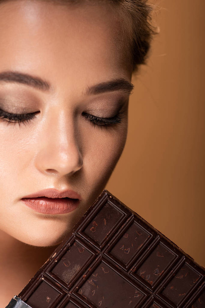 mooie jonge vrouw met gesloten ogen houden chocoladereep geïsoleerd op beige - Foto, afbeelding