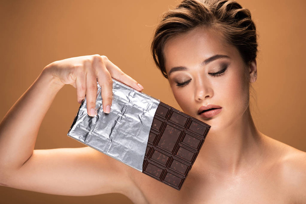 beautiful young naked woman with closed eyes holding chocolate bar in silver foil isolated on beige - Foto, afbeelding