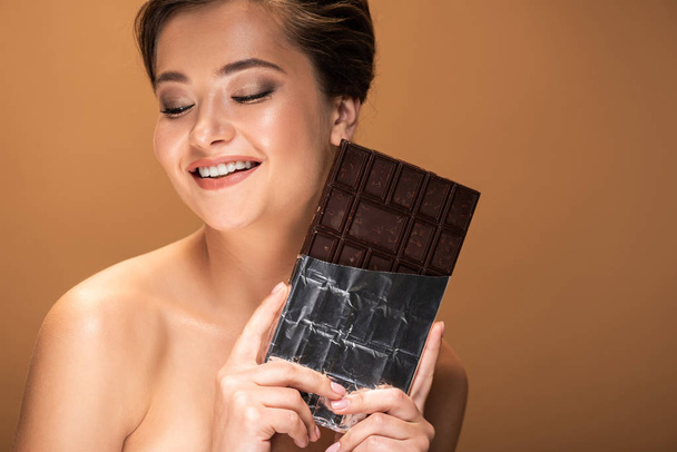 όμορφη χαμογελαστή νεαρή γυμνή γυναίκα κρατώντας μπάρα σοκολάτας σε ασημένιο φύλλο απομονωμένο σε μπεζ - Φωτογραφία, εικόνα