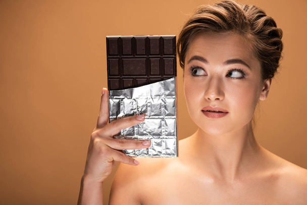 mooie jonge naakte vrouw kijken naar chocolade bar in zilver folie geïsoleerd op beige - Foto, afbeelding