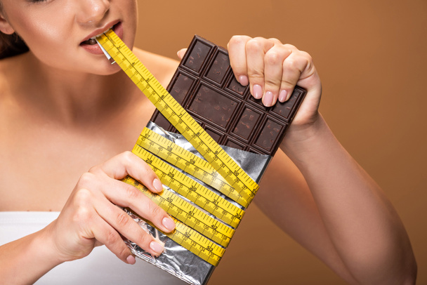 口に黄色の測定テープを持つ若い女性の部分的なビューとベージュで分離されたチョコレートバー - 写真・画像