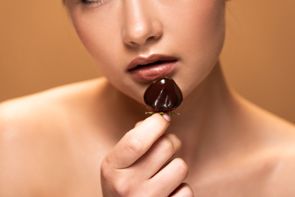 обрезанный вид красивой обнаженной женщины, держащей клубнику в расплавленном шоколаде, изолированном на бежевом
 - Фото, изображение