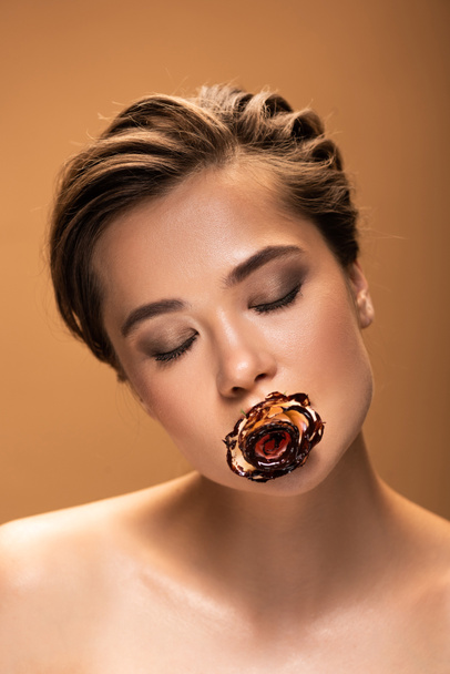 目を閉じた裸の女性は、ベージュに隔離された溶けたチョコレートで覆われた口の中でバラを保持しています - 写真・画像