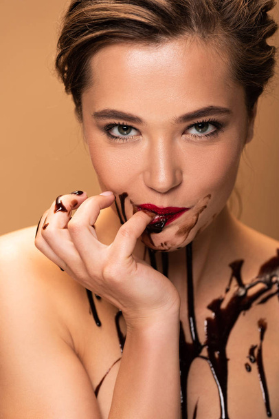 γυμνή γυναίκα με κόκκινα χείλη και σοκολάτα σταγόνες στο δέρμα γλείφει το δάχτυλο απομονωθεί σε μπεζ - Φωτογραφία, εικόνα
