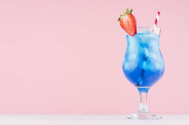 Tropical beach alcohol blue drink  - curacao liquor, strawberry slice, ice, straw in misted glass on pink background, white wood board. - Φωτογραφία, εικόνα