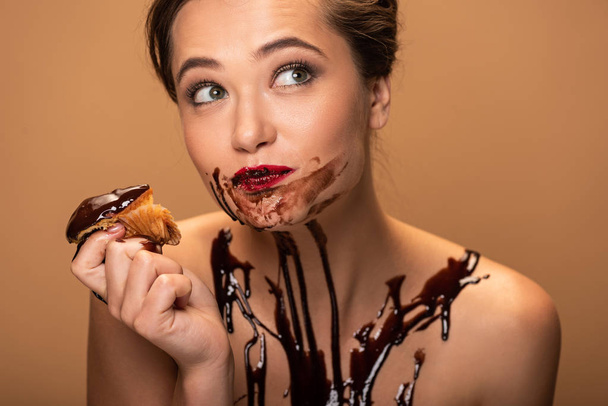 beautiful dreamy naked woman with red lips and chocolate spills on skin eating muffin isolated on beige - Photo, Image