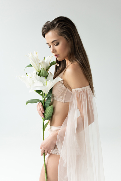 tender young woman in beige lingerie and mesh sleeves looking at lilies isolated on white - Fotoğraf, Görsel