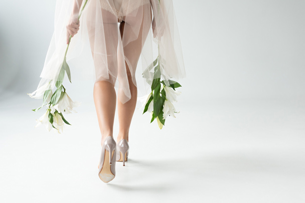 Rückansicht einer jungen Frau in beiger Dessous und Netzärmeln, die mit Lilien auf weißem Hintergrund spazieren geht - Foto, Bild