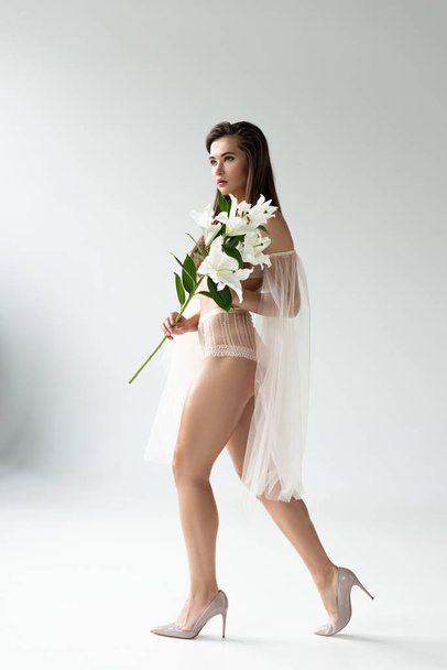 side view of tender young woman in beige lingerie and mesh sleeves walking with white lilies - Fotó, kép