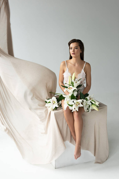 tender young woman in white lacy bodysuit sitting on cube near beige curtain with lilies isolated on white - Foto, Imagem