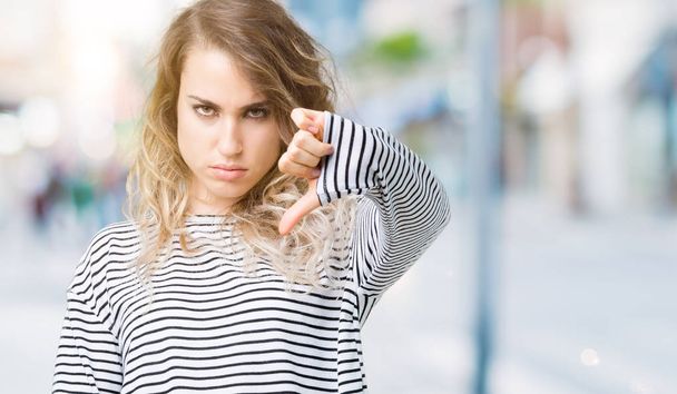 美しい若い金髪女性の不幸と怒りの拒否反応を示さずとジェスチャーを親指で否定的な分離の背景にストライプのセーターを着ています。不適切な表現. - 写真・画像
