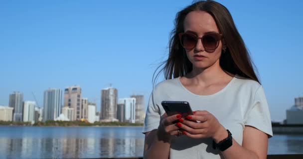 Hermosa chica con el pelo largo en gafas de sol usando aplicación de teléfono inteligente en el muelle del río puesta de sol cerca de 4K
. - Metraje, vídeo
