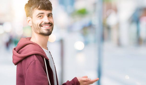 Jonge knappe man over geïsoleerde achtergrond wijst naar de kant met de hand en open palm, presenteren ad glimlachend gelukkig en vol vertrouwen - Foto, afbeelding