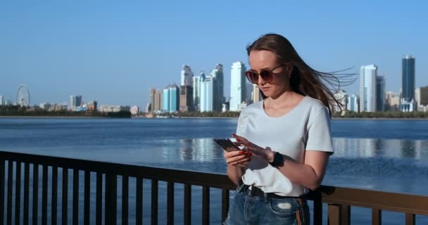 Krásná dívka s dlouhými vlasy v slunečních brýlích, která používá smartphone při západu slunce na nábřeží poblíž 4k. - Záběry, video