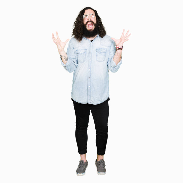 Homem jovem hipster com cabelos longos e barba vestindo óculos loucos e loucos gritando e gritando com expressão agressiva e braços levantados. Conceito de frustração
. - Foto, Imagem