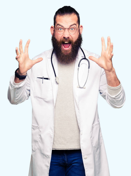 Young blond doctor man with beard wearing medical coat celebrating crazy and amazed for success with arms raised and open eyes screaming excited. Winner concept - Photo, Image