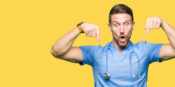 分離上医療制服を着てハンサムな医者男ダウン表示広告、驚いた顔、口を開けて指でポインティングを背景します。 - 写真・画像