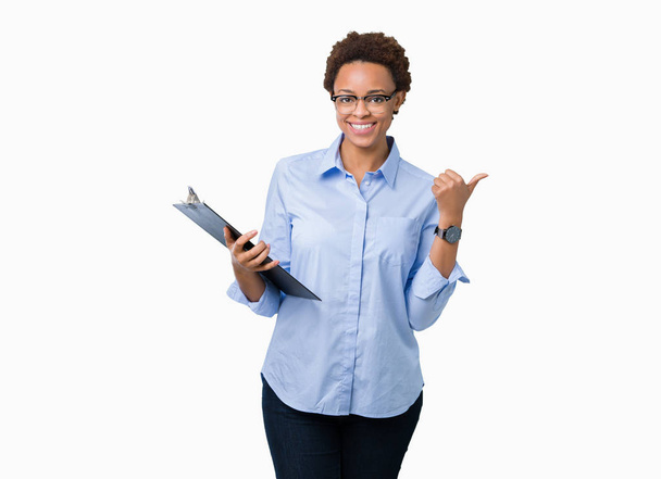junge afrikanisch-amerikanische Geschäftsfrau hält Klemmbrett über isoliertem Hintergrund und zeigt mit dem Daumen zur Seite mit glücklichem Lächeln - Foto, Bild