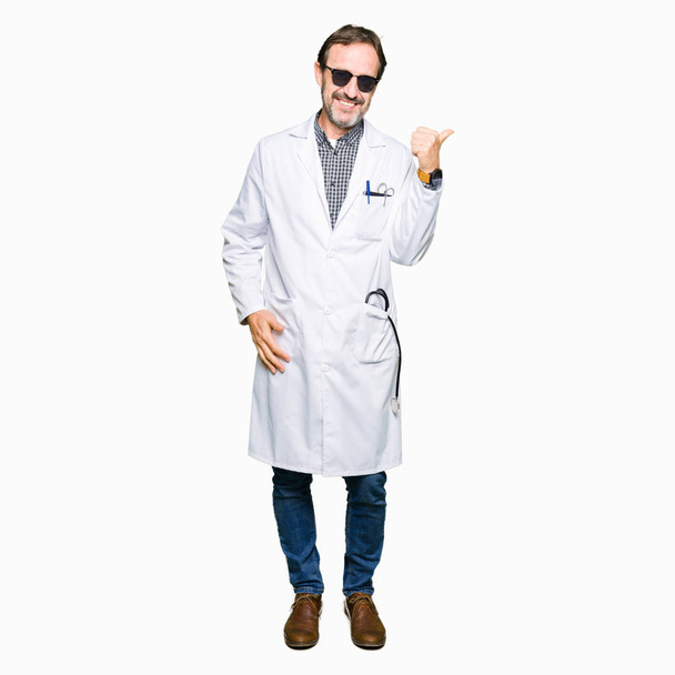 Handsome middle age doctor man wearing sunglasses smiling with happy face looking and pointing to the side with thumb up. - Photo, Image