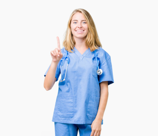 Linda jovem médica mulher vestindo uniforme médico sobre fundo isolado mostrando e apontando para cima com o dedo número um, sorrindo confiante e feliz
. - Foto, Imagem