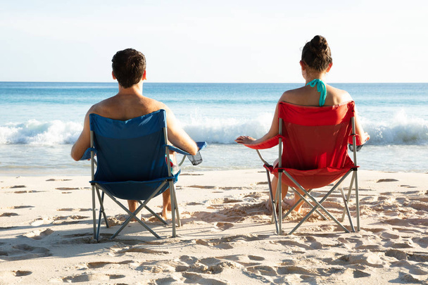 Rear View Of Young Couple Sitting On Chairs Looking At Blue Sea On Beach - Фото, изображение