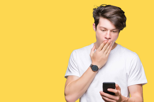 Young man using smartphone over isolated background cover mouth with hand shocked with shame for mistake, expression of fear, scared in silence, secret concept - Zdjęcie, obraz