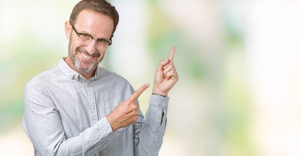 Όμορφος μέση ηλικία κομψό ανώτερος άνθρωπος φορώντας γυαλιά πέρα από το απομονωμένο υπόβαθρο χαμογελώντας και εξετάζοντας τη φωτογραφική μηχανή κατάδειξης με δύο χέρια και τα δάχτυλα στο πλάι. - Φωτογραφία, εικόνα