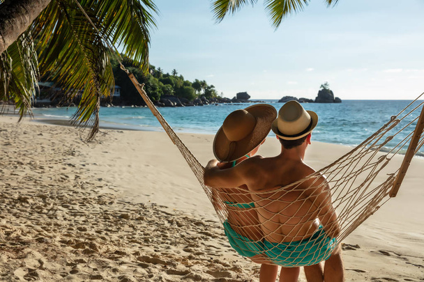 Молода пара носіння капелюх сидячи на гамаку разом дивлячись на ідилічне море на пляжі - Фото, зображення