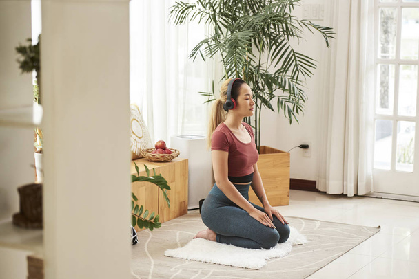 Młoda Azjatka w słuchawkach siedzi na puszystym dywanie i medytuje - Zdjęcie, obraz