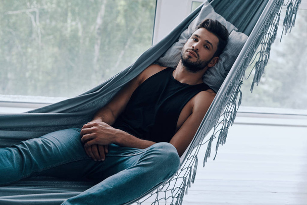 Handsome young man in casual clothing looking at camera while lying in hammock indoors - Foto, immagini