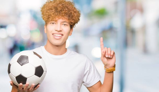 Młody przystojny mężczyzna trzyma piłkę nożną piłki nożnej zaskoczony pomysłem lub pytaniem wskazującym palcem z szczęśliwą twarzą, numer jeden - Zdjęcie, obraz