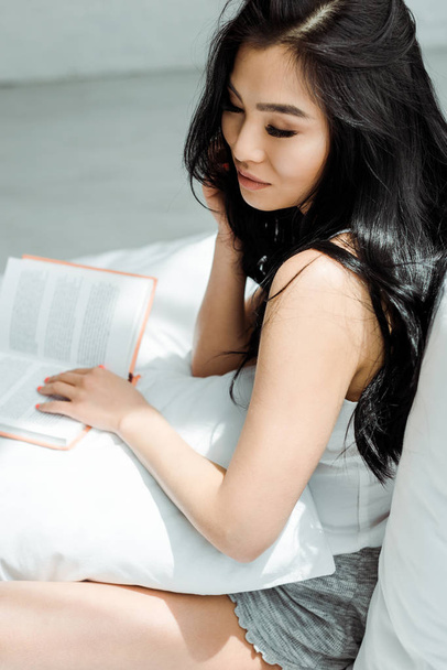 αρκετά ασιατικό κορίτσι αγγίζοντας τα μαλλιά και κρατώντας το βιβλίο ενώ κάθεται στο κρεβάτι  - Φωτογραφία, εικόνα